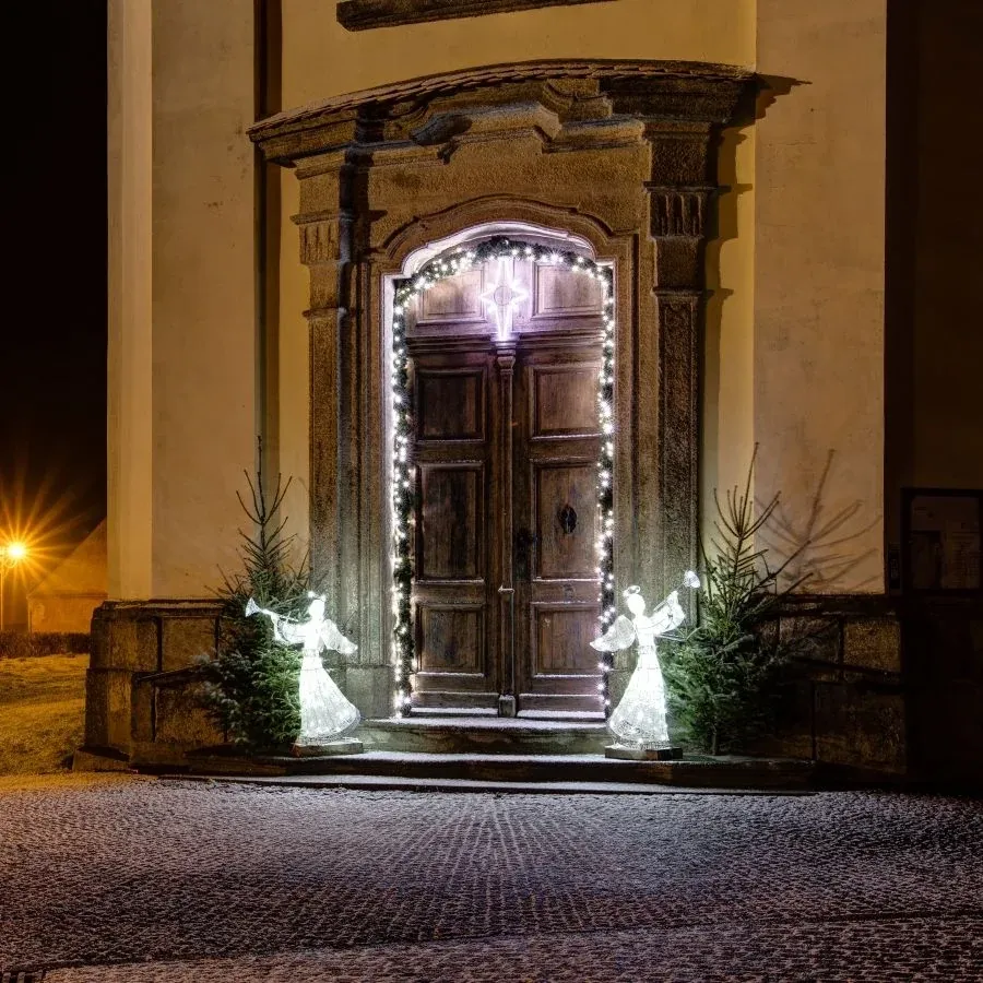 Zwei eisweiße LED-Engel mit Trompete an einer Barocktür mit Weihnachtsbäumchen, nächtliche Aufnahme des Eingangs